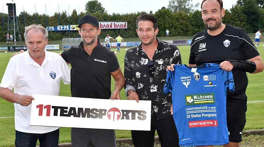 Vier Männer auf einem Fußballfeld präsentieren ein Teamsport-Schild und ein Fußballtrikot.