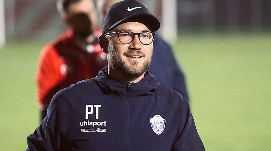 Mann im Sportanzug und Cap auf einem Fußballplatz, abends beleuchtet, lächelnd in die Kamera schauend.