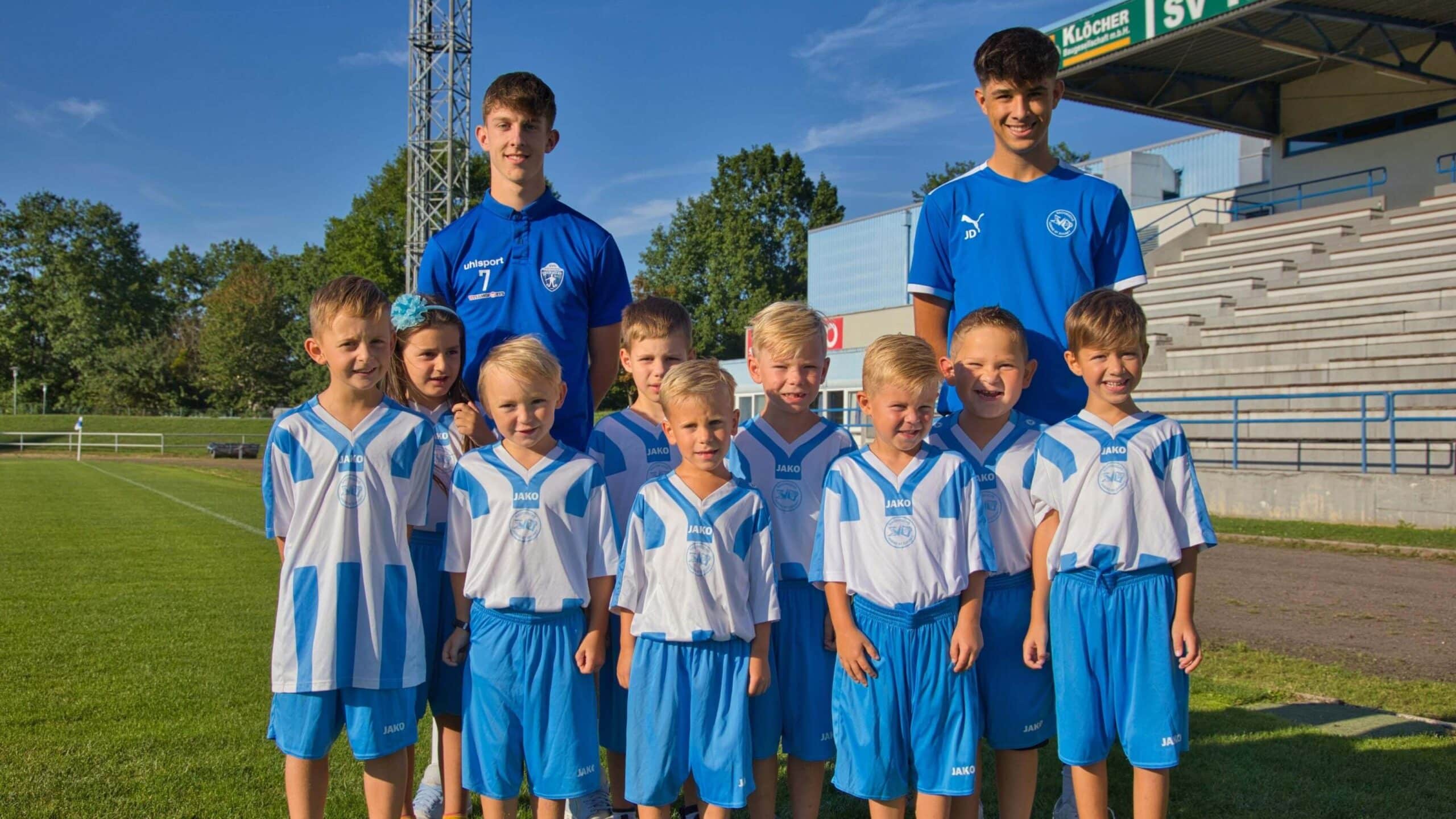Jugendfußballteam in blauen und weißen Trikots posiert mit zwei Trainern auf einem Sportplatz bei sonnigem Wetter.
