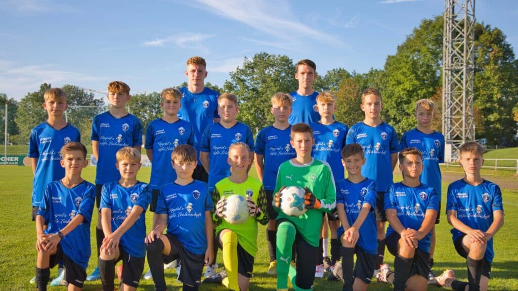 Jugendfußball-Team in blauen Trikots posiert auf dem Fußballfeld für ein Mannschaftsfoto bei Sonnenschein.