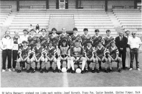 Fußballmannschaft posiert für ein Gruppenfoto im Stadion, Spieler in gestreiften Trikots, Trainer und Teammitglieder.