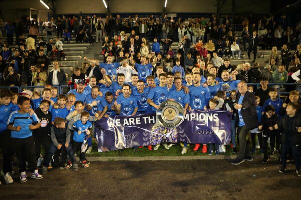 Fußballteam feiert Meisterschaftssieg mit Fans und Trophäe auf dem Spielfeld, Gruppenfoto abends im Stadion.