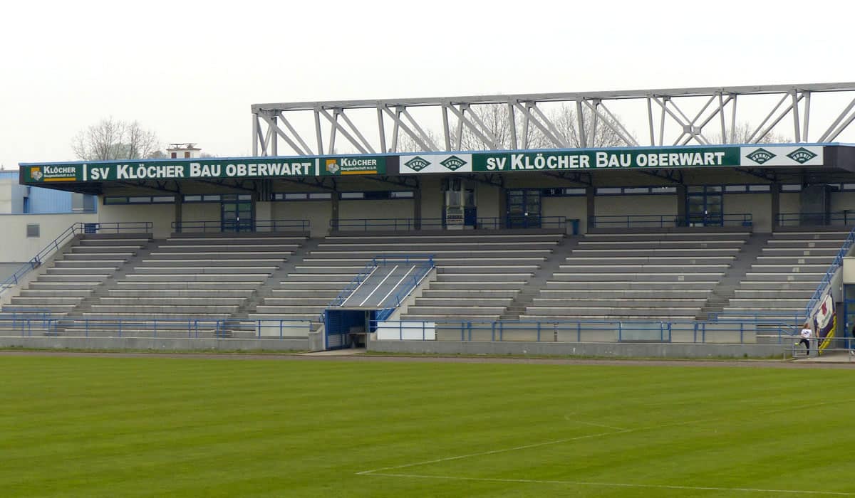 Leere Fußballtribüne im SV Klöcher Bau Oberwart Stadion, mit grüner Werbetafel und Sitzplätzen.