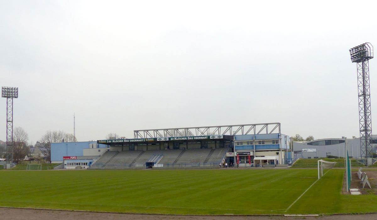 Fußballstadion mit Tribünen und Flutlichtmasten an einem bewölkten Tag, SV Klöcher Bau Oberwart.