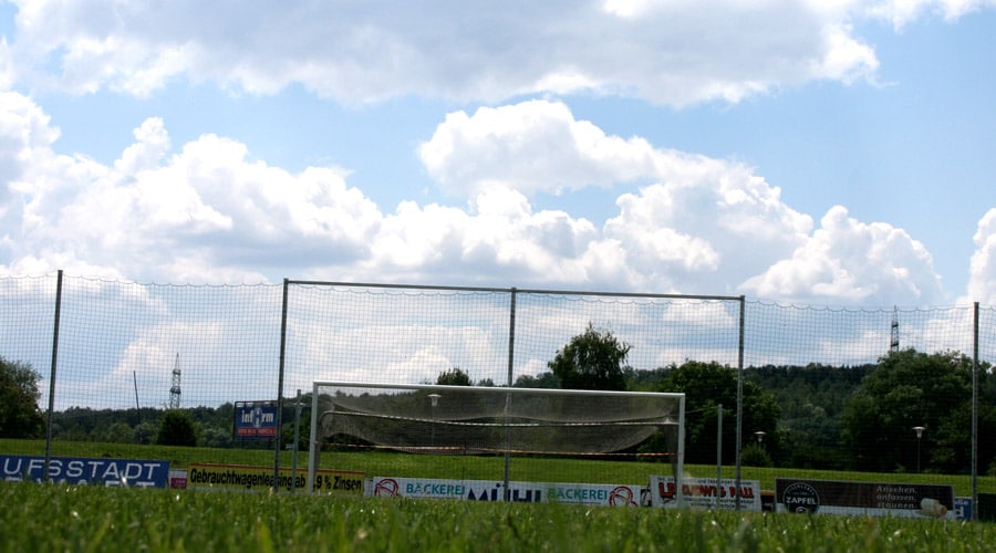 Fußballtor auf grünem Spielfeld unter bewölktem Himmel, umgeben von Bäumen und Reklametafeln.
