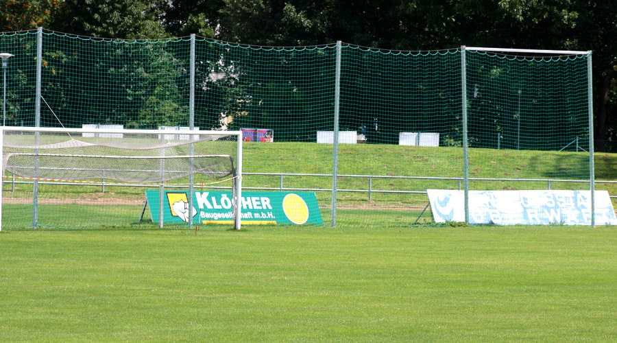 Fußballtor auf einem grünen Sportplatz mit Werbebannern und Schutznetz im Hintergrund.