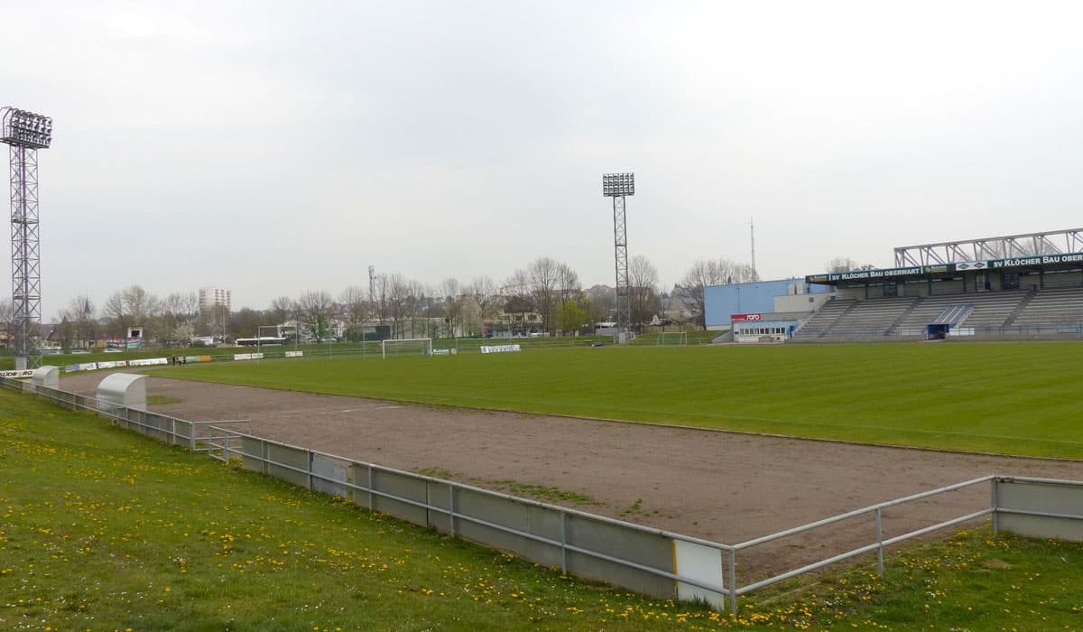 Grünes Fußballstadion mit leeren Tribünen und Flutlichtmasten an einem bewölkten Tag. Ideal für Sportveranstaltungen.