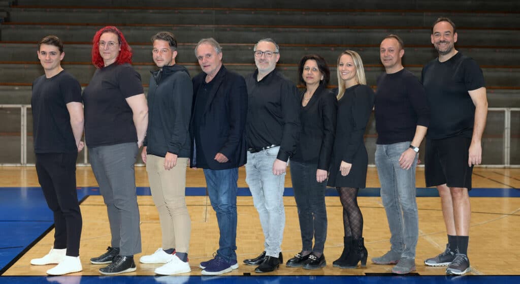 Acht Personen stehen in einer Turnhalle in einer Linie und tragen überwiegend schwarze Kleidung.