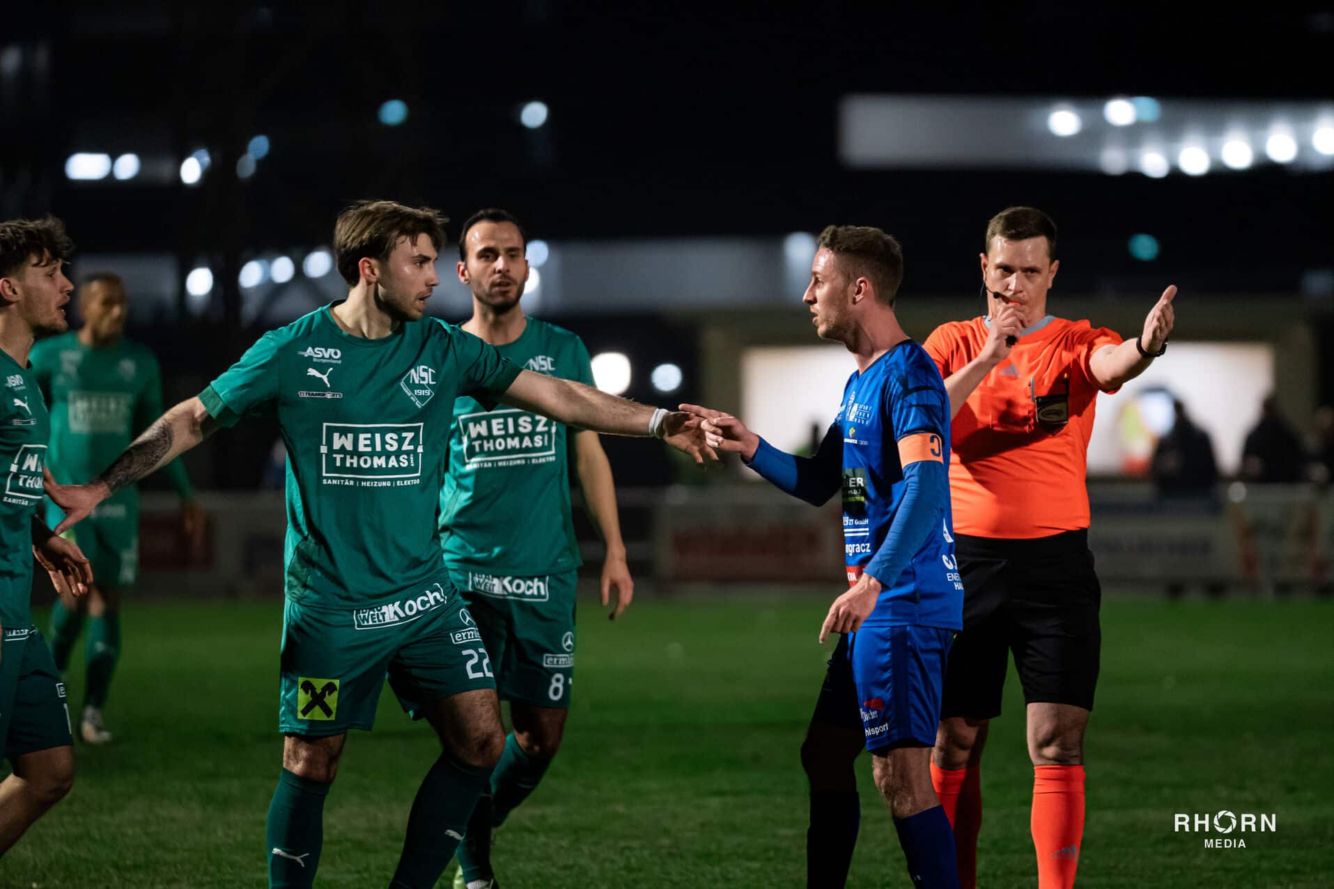 Schiedsrichter trennt Spieler in grün und blau bei einem nächtlichen Fußballspiel auf dem Spielfeld.