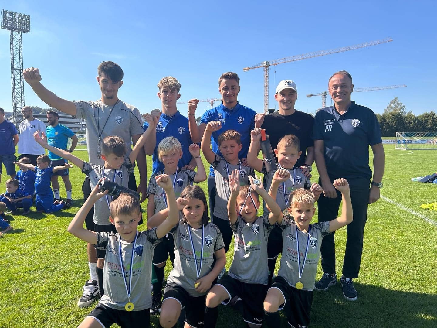 Kinder-Fußballteam feiert mit Trainern auf dem Spielfeld nach einem erfolgreichen Turnier, bei sonnigem Wetter.