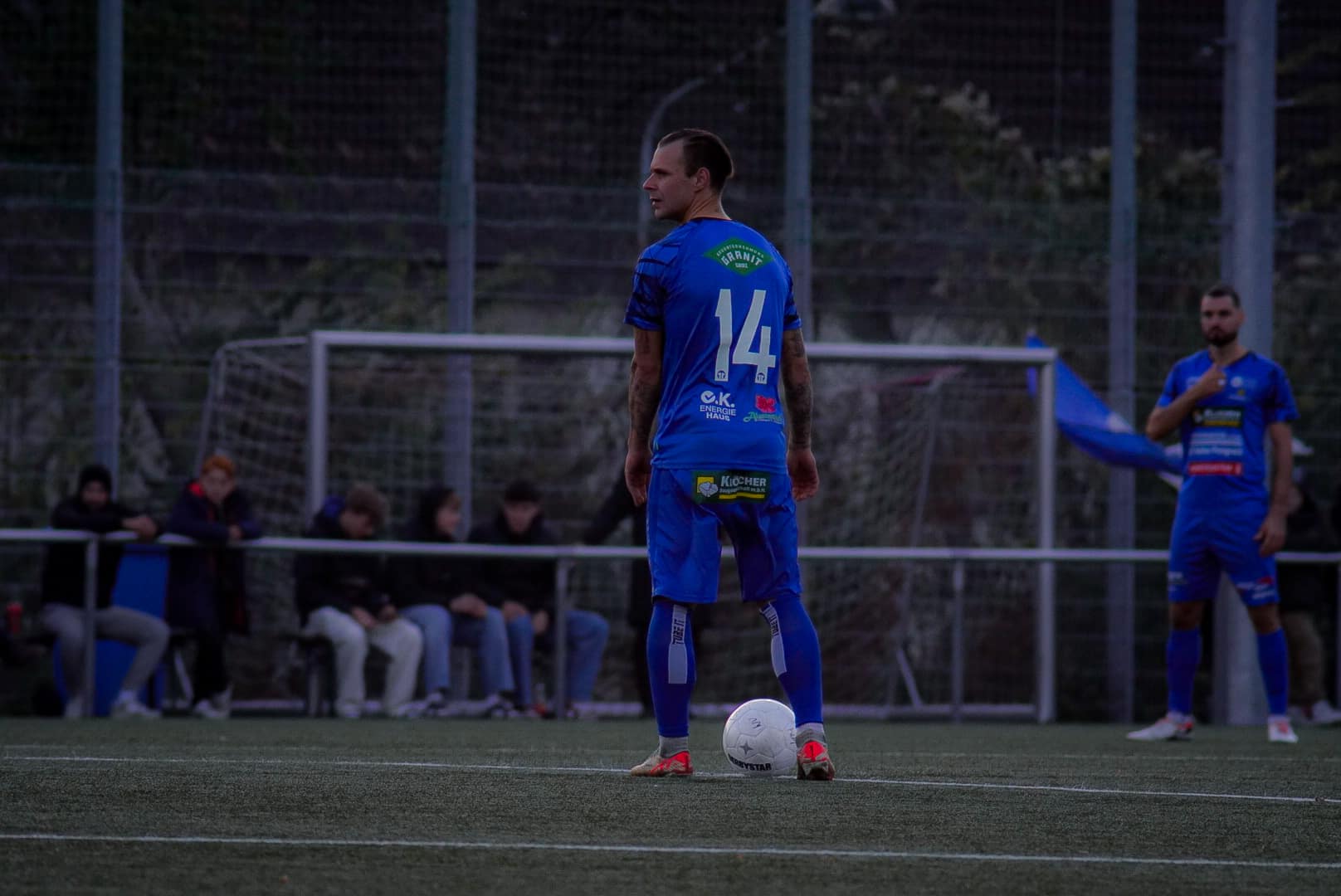 Fußballspieler in blauem Trikot mit Nummer 14 bereitet sich auf einen Freistoß im Abendlicht vor.