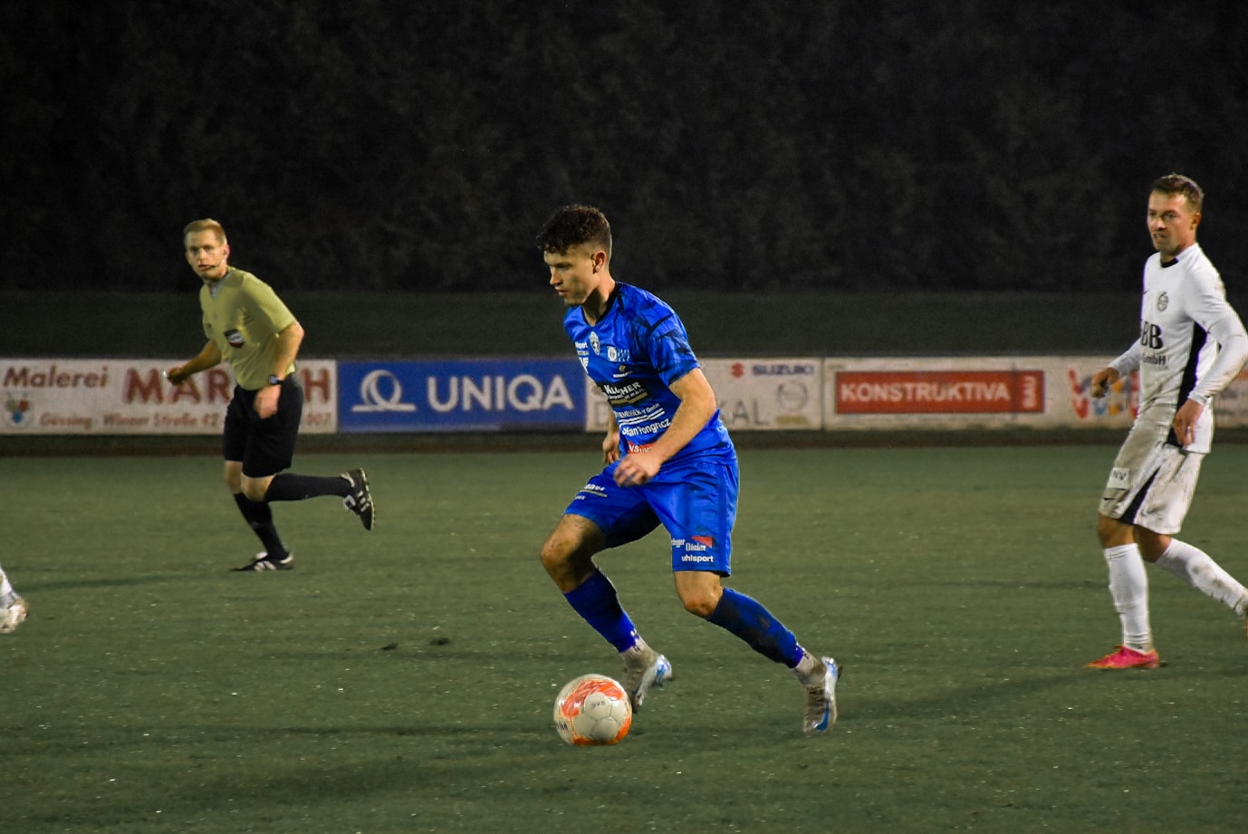 Fußballspieler in blauem Trikot dribbelt den Ball auf dem Spielfeld, verfolgt von Gegnern und beobachtet vom Schiedsrichter.