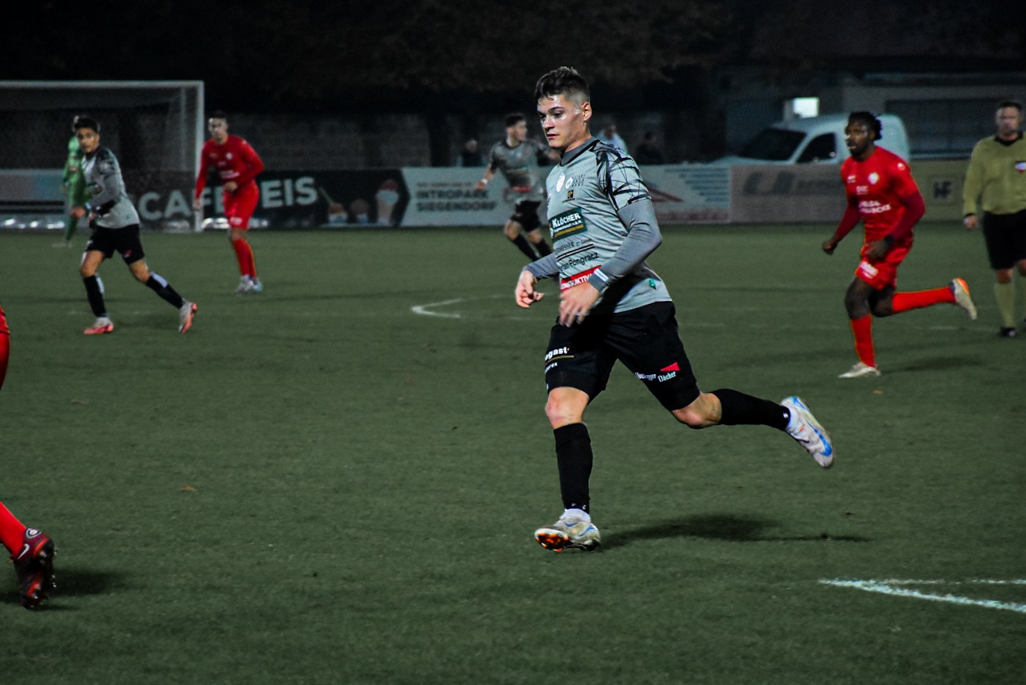 Fußballspieler im grauen Trikot läuft über das Spielfeld während eines Abends, umgeben von Spielern in roten Trikots.