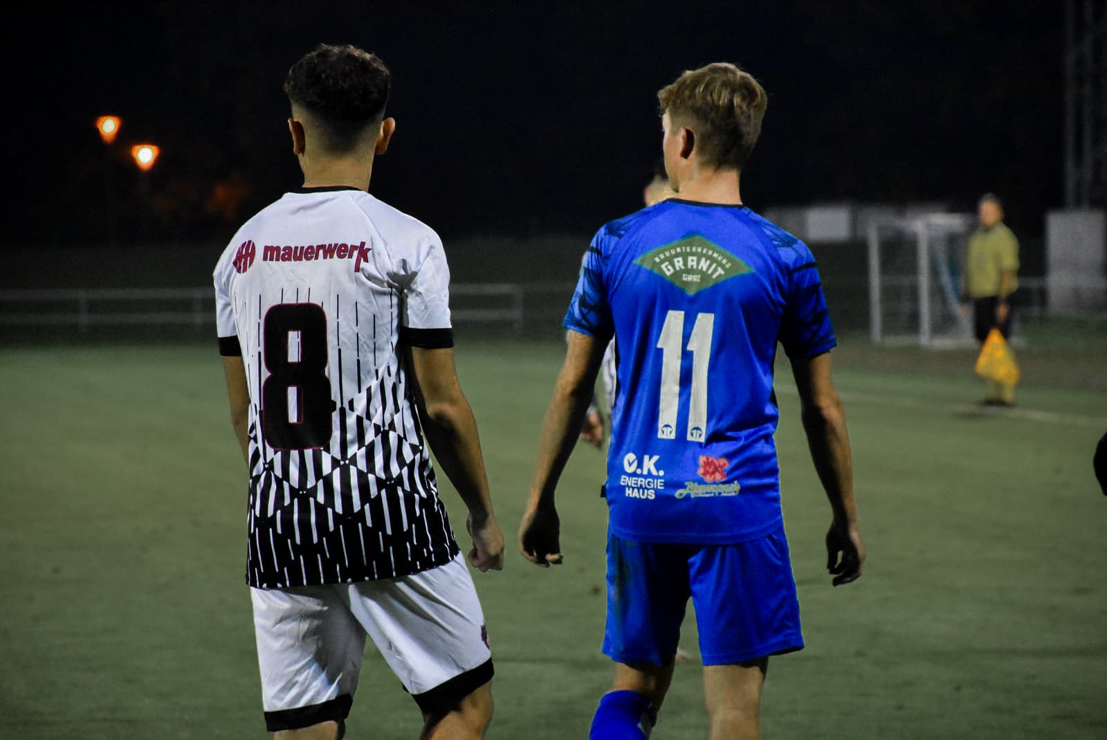 Zwei Fußballspieler im Trikot auf dem Spielfeld bei Nacht, Rückenansicht.