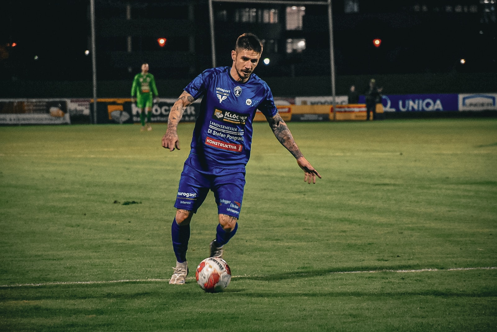 Fußballspieler im blauen Trikot kontrolliert den Ball während eines Nachtspiels auf dem Rasenplatz.