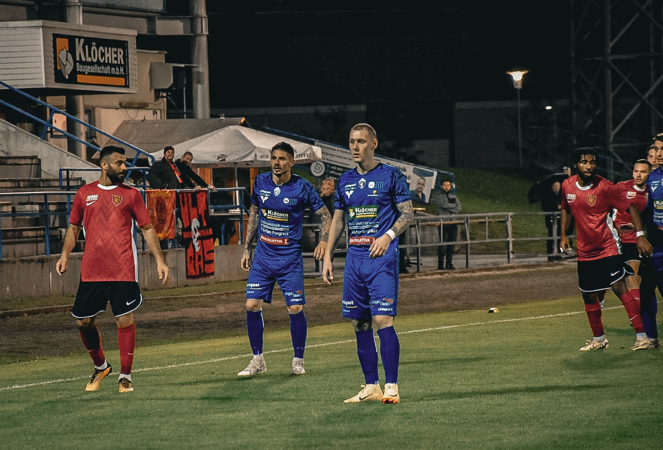 Fußballspiel bei Nacht, zwei Teams in Rot und Blau, Spieler im Fokus, Stadion im Hintergrund, spannendes Match.