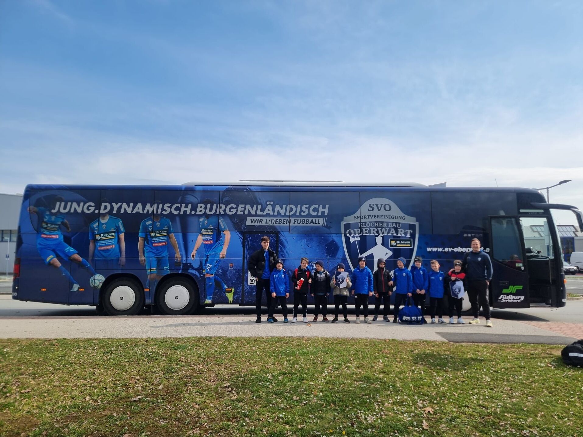 Jugendfußballteam vor bemaltem Vereinsbus der Sportvereinigung Oberwart, bereit für ein spannendes Match.