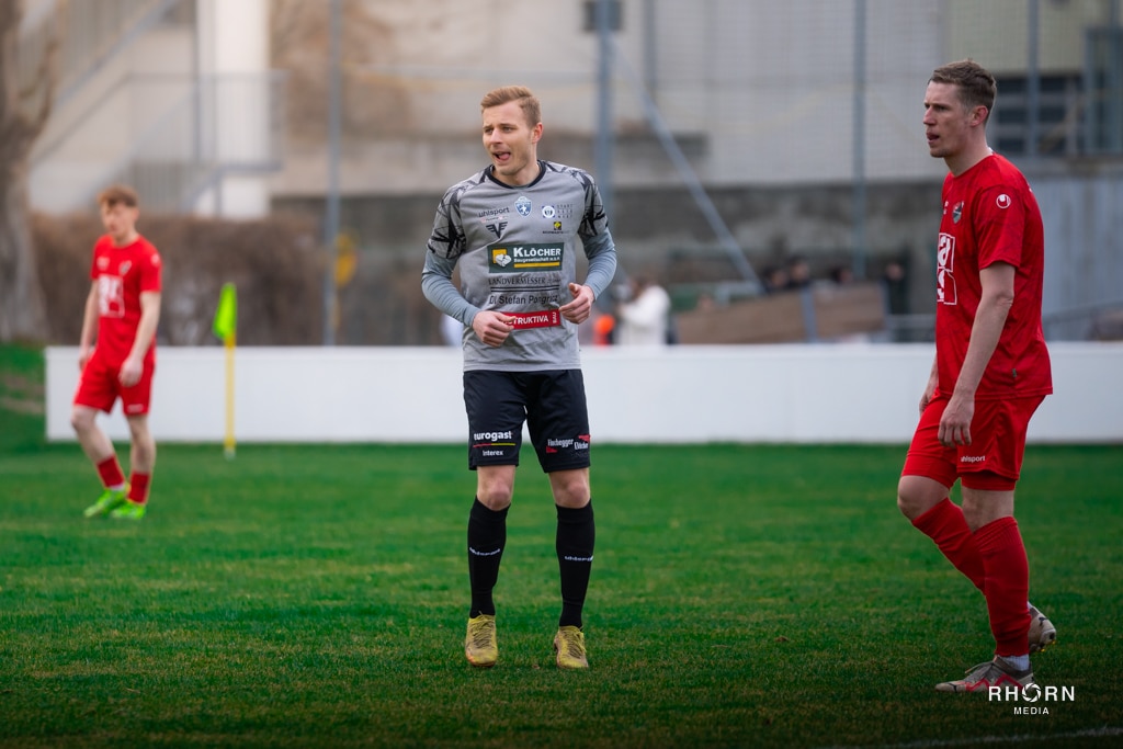Fußballspieler auf dem Feld während eines Spiels, einer trägt graues Trikot, andere in roten Trikots, grüner Rasen.