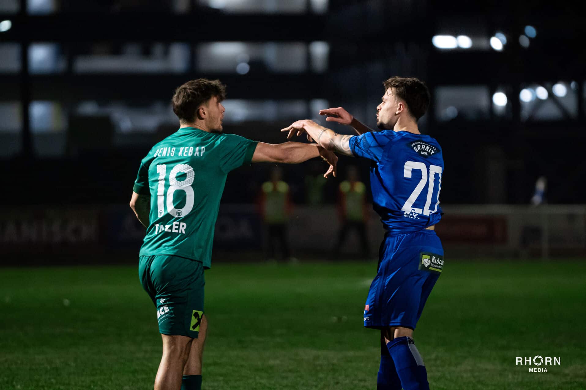 Zwei Fußballspieler diskutieren intensiv während eines Spiels bei Nacht, Trikots mit Nummern 18 und 20.