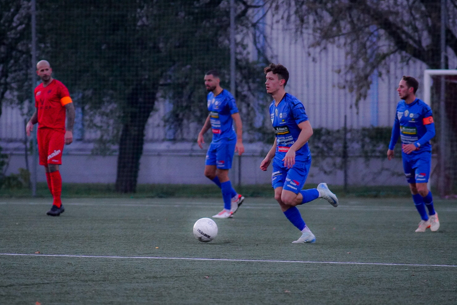 Fußballspieler in blauer und roter Uniform im Spiel auf grünem Feld, während sie den Ball kontrollieren.