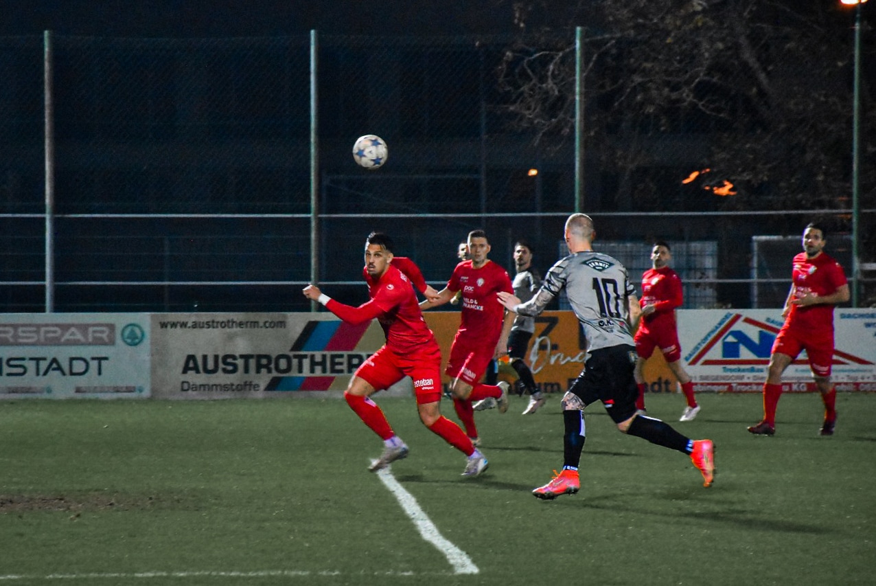 Fußballspiel bei Nacht: Spieler in roten Trikots und einem in Grau verfolgen den Ball auf einem beleuchteten Spielfeld.