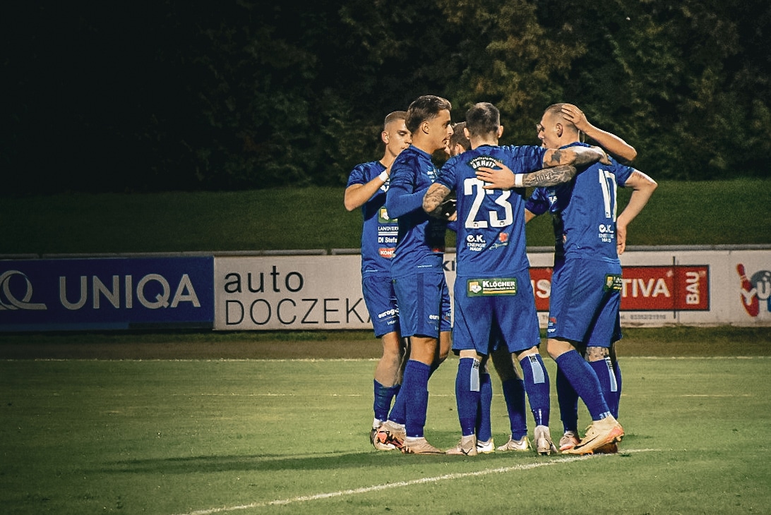 Fußballmannschaft in blauen Trikots feiert gemeinsam auf dem Spielfeld nach einem Torerfolg bei einem Abendspiel.