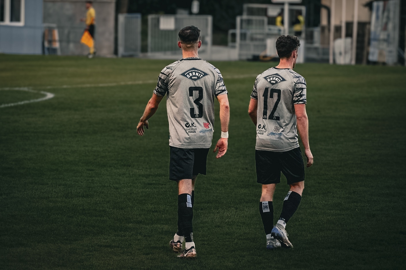 Zwei Fußballspieler in grauen Trikots mit den Nummern 3 und 12 auf einem Spielfeld, von hinten gesehen.