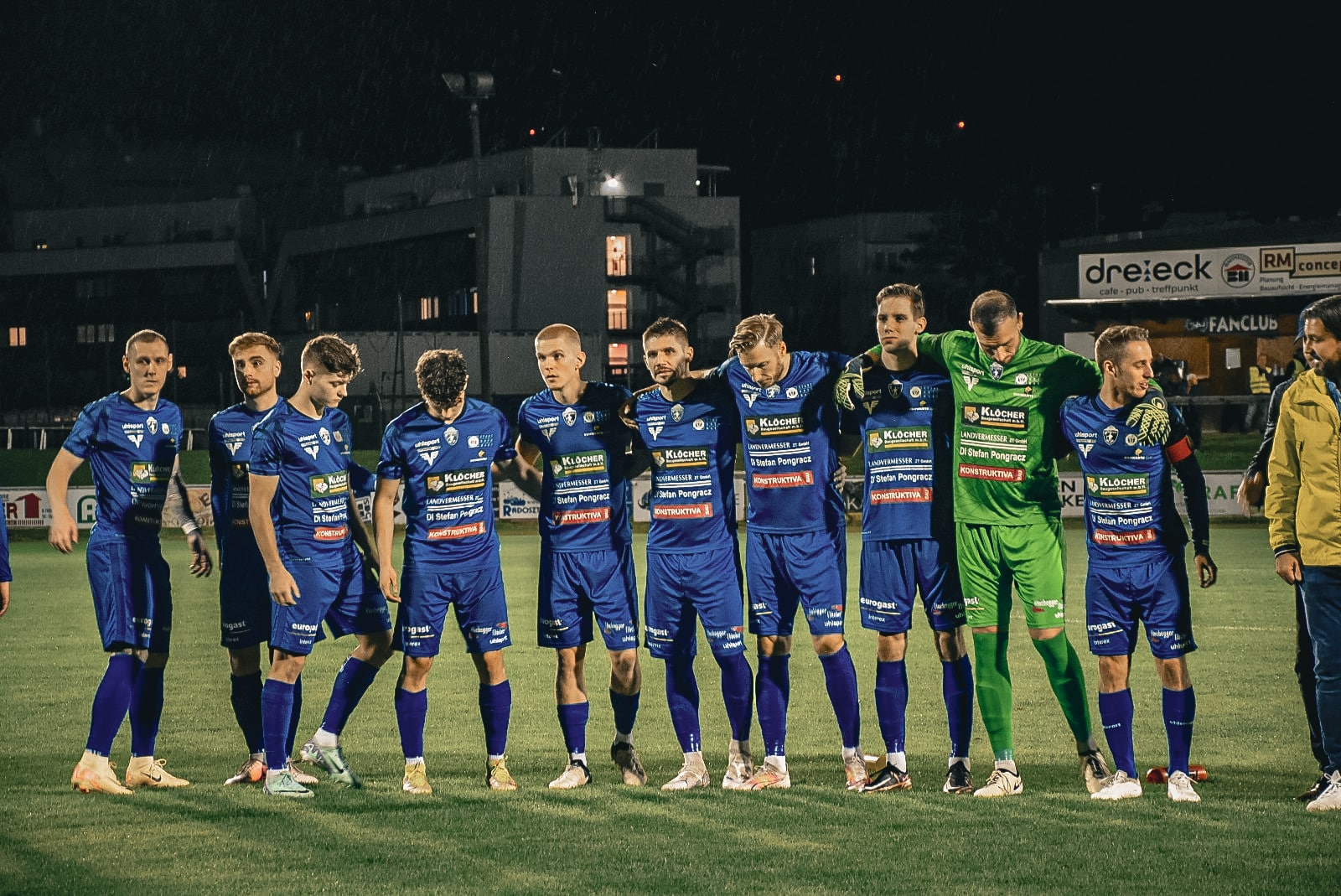 Fußballmannschaft in blauen Trikots in einer Linie auf dem Spielfeld bei Nacht. Teamgeist und Zusammenhalt stärken.