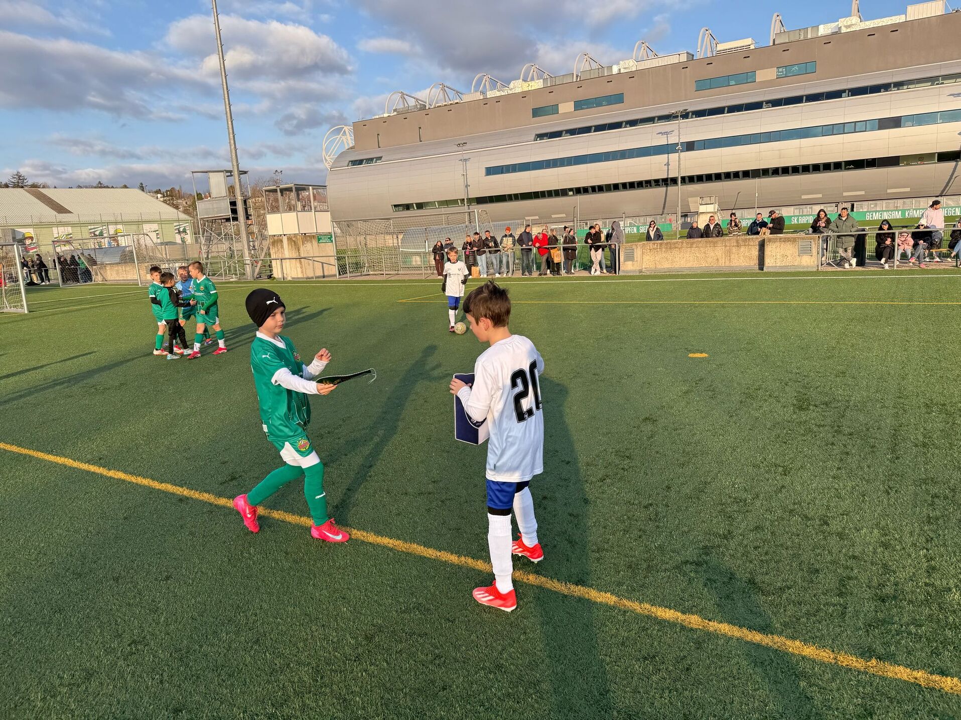 Kinder spielen Fußball auf einem Sportplatz, Stadion im Hintergrund, sonniger Tag, Zuschauer beobachten das Spiel.