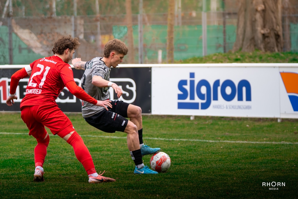 Fußballspiel, zwei Spieler im Zweikampf um den Ball, vor einer Werbebande auf dem Spielfeld.
