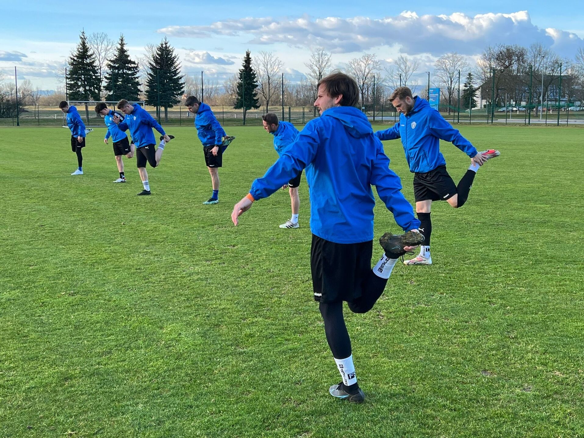 Fußballspieler in blauen Jacken dehnen sich auf grünem Rasen bei sonnigem Wetter.