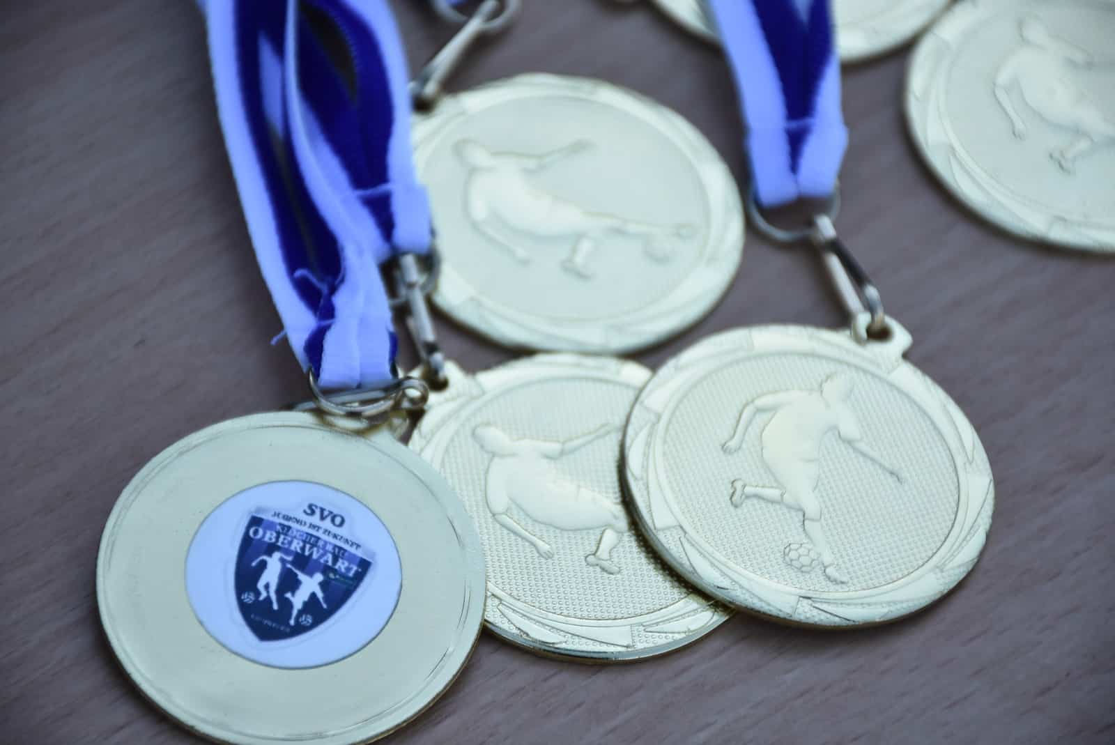 Goldene Medaillen mit blau-weißen Bändern für ein Fußballturnier auf Holzuntergrund.
