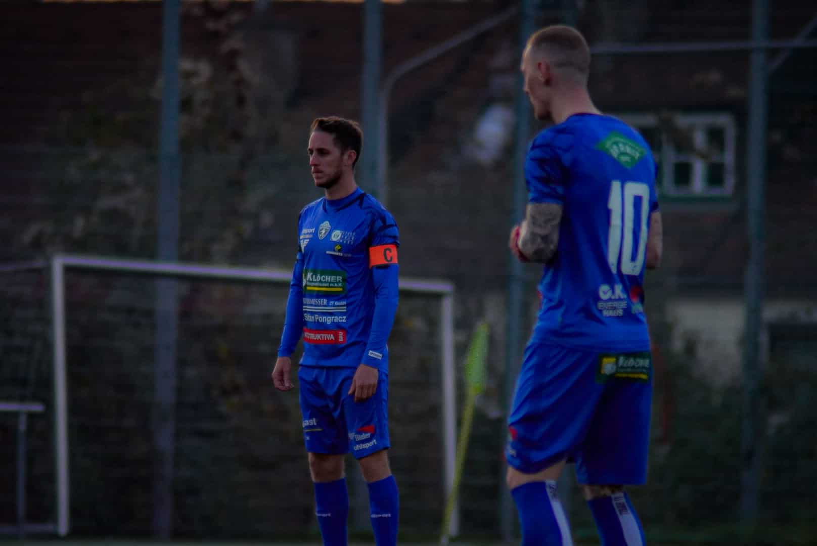Zwei Fußballspieler in blauen Trikots bereiten sich auf ein Spiel vor, einer mit Kapitänsbinde, im Hintergrund ein Tor.