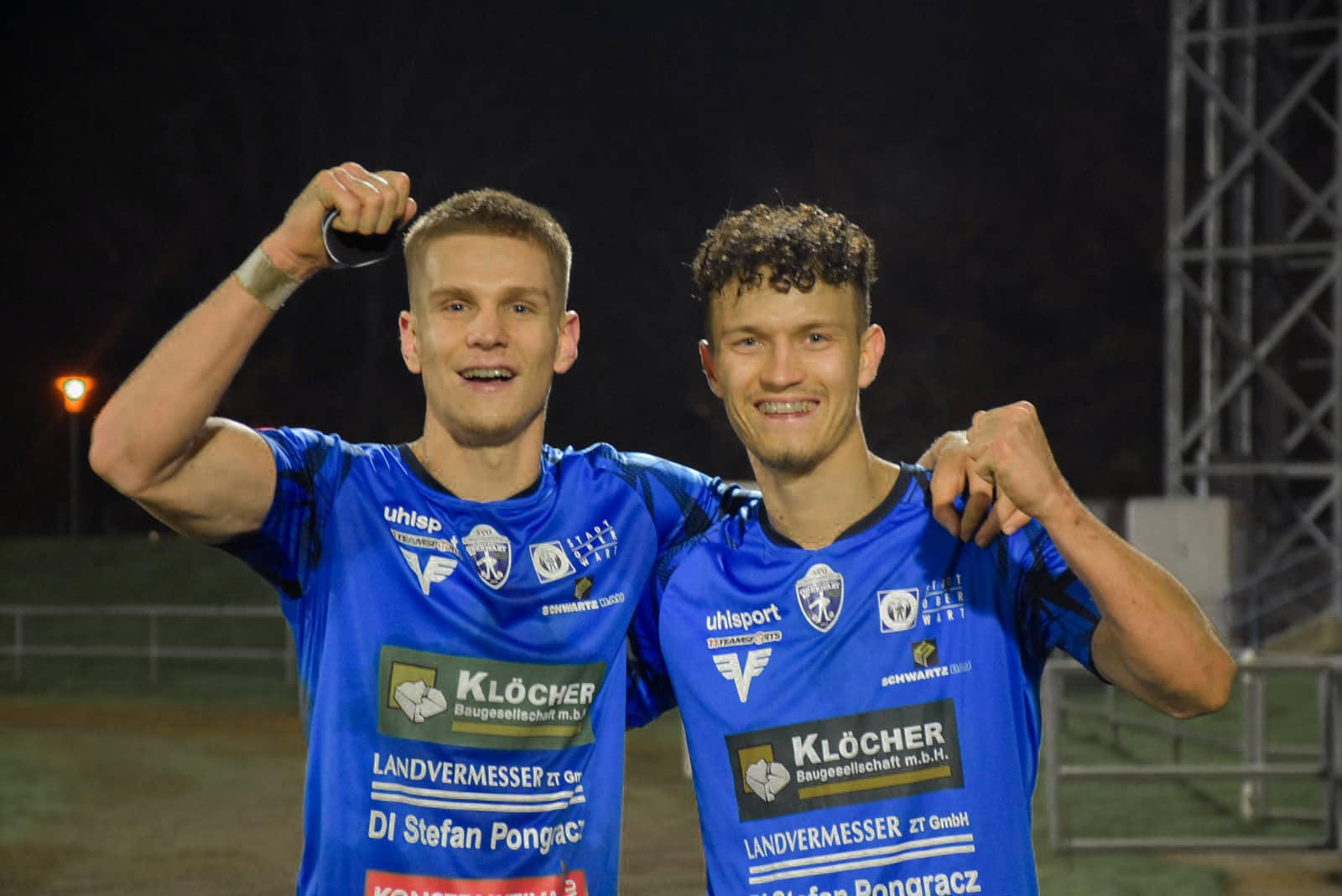 Zwei Fußballspieler in blauen Trikots jubeln auf dem Spielfeld unter Flutlicht. Erfolgreicher Sieg beim Abendspiel.