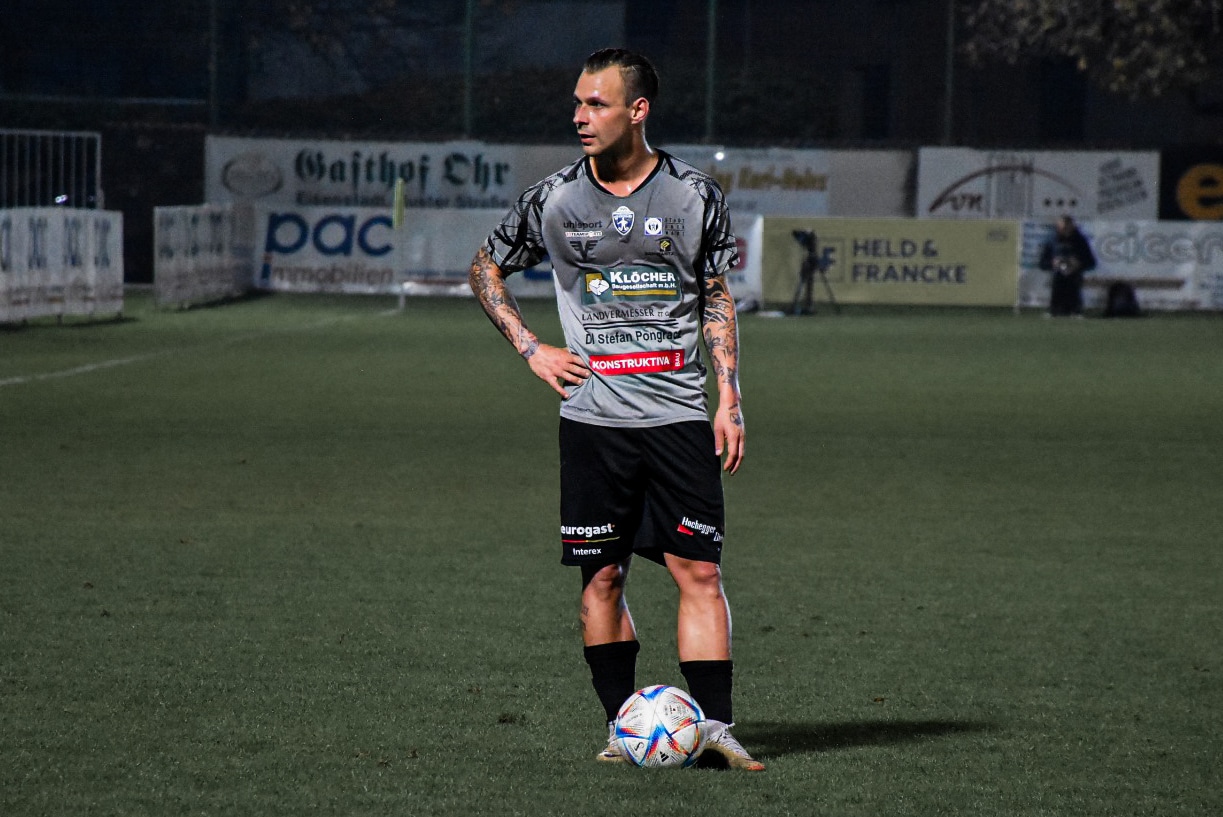 Fußballspieler in grauem Trikot, bereit für den Freistoß auf einem beleuchteten Spielfeld in der Nacht.