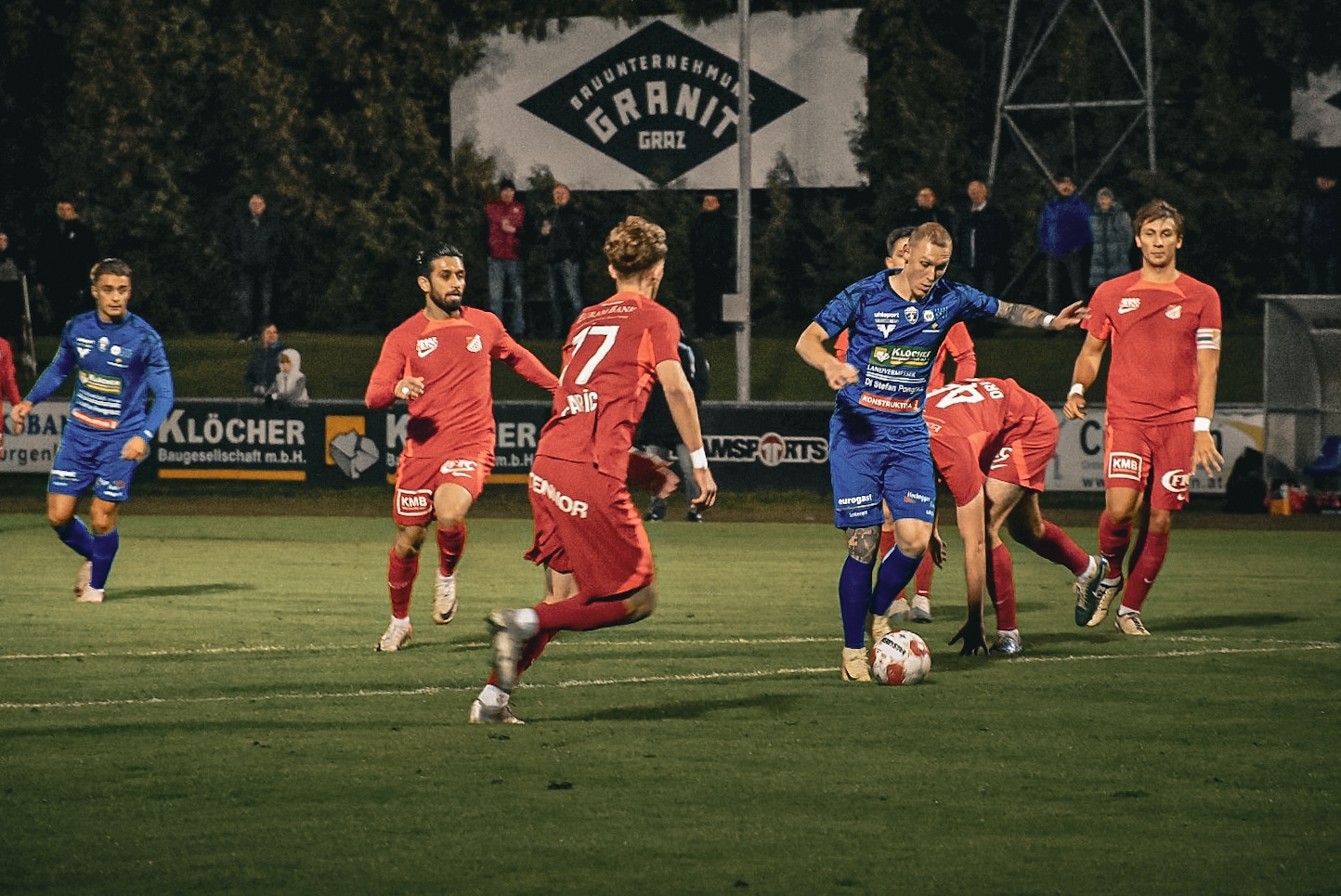 Fußballspiel bei Nacht: Spieler in blauem Trikot dribbelt den Ball, umgeben von Spielern in roten Trikots auf dem Feld.