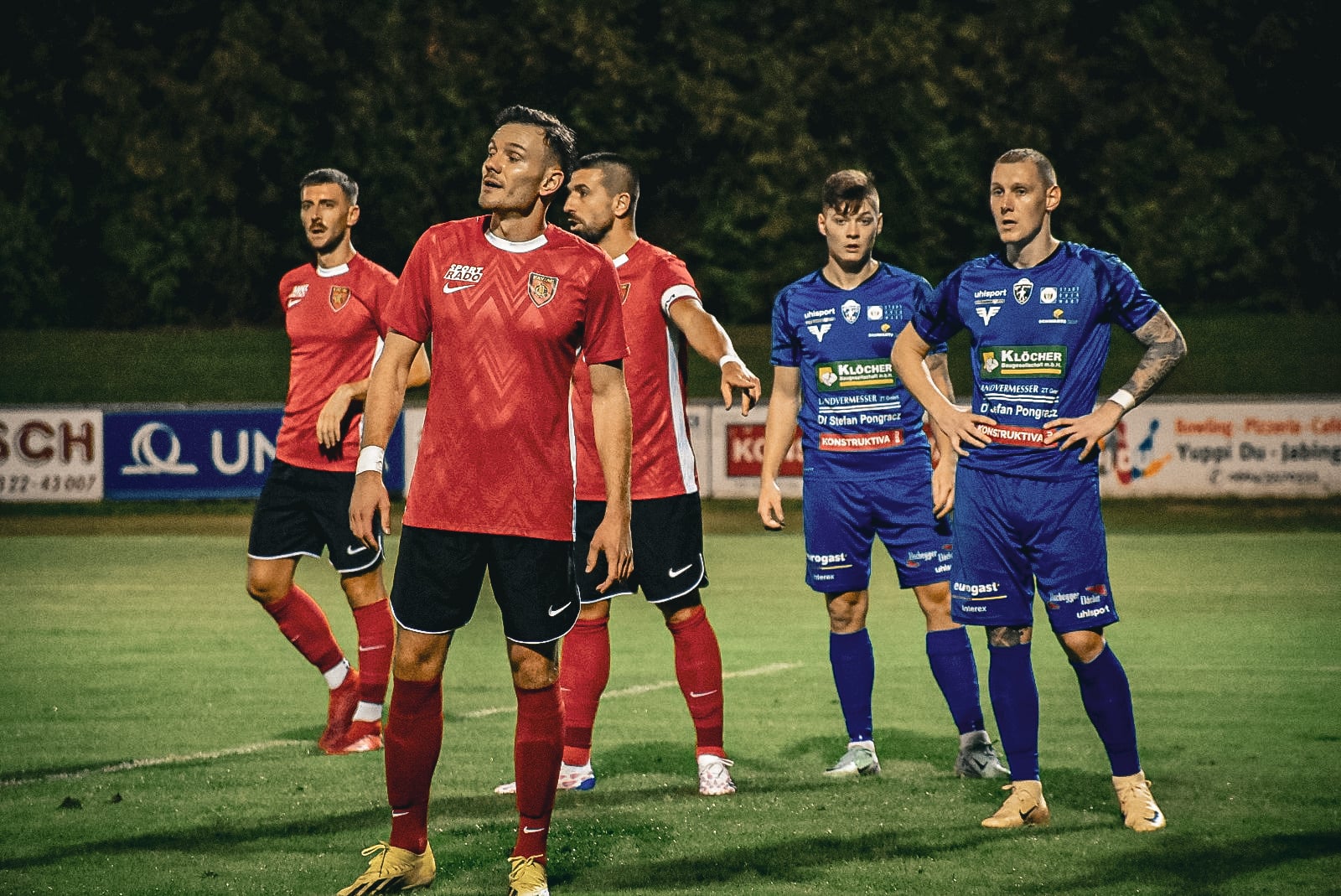 Fußballspiel, rote gegen blaue Trikots, Spieler in Erwartung auf dem Spielfeld, abends im Freien.