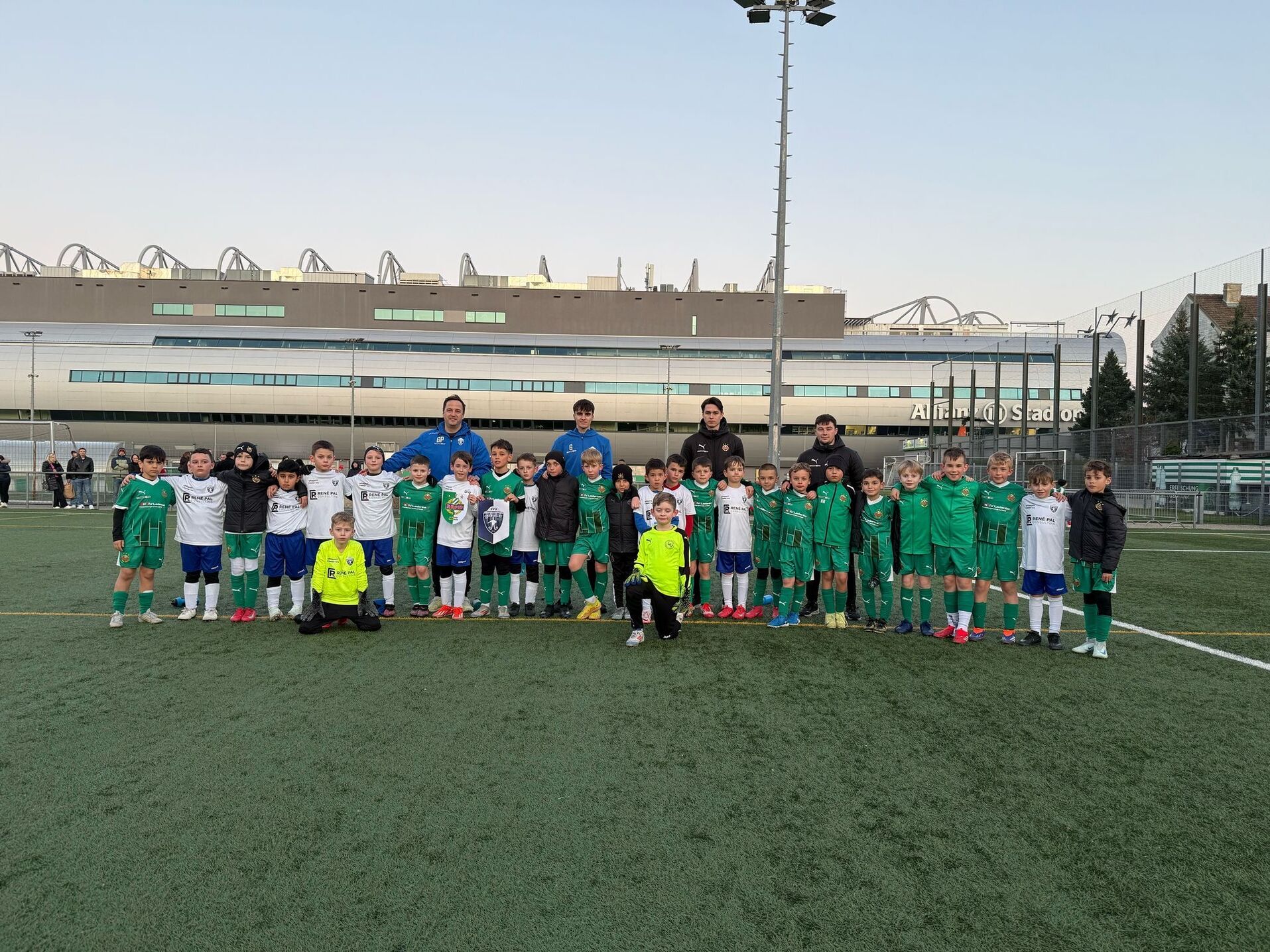 Gruppe von jungen Fußballspielern auf einem Sportplatz vor einem Stadion, lächelnd und bereit für das Spiel.