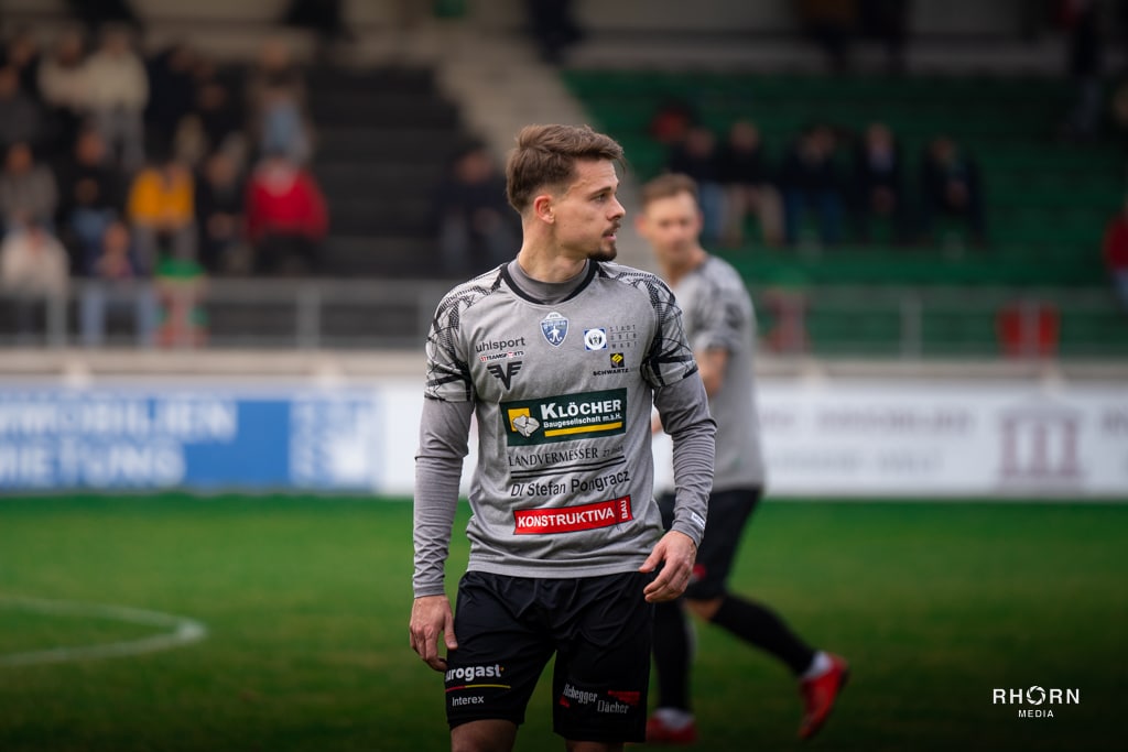 Fußballspieler im grauen Trikot auf dem Spielfeld, konzentriert in Aktion, Zuschauer im Hintergrund.