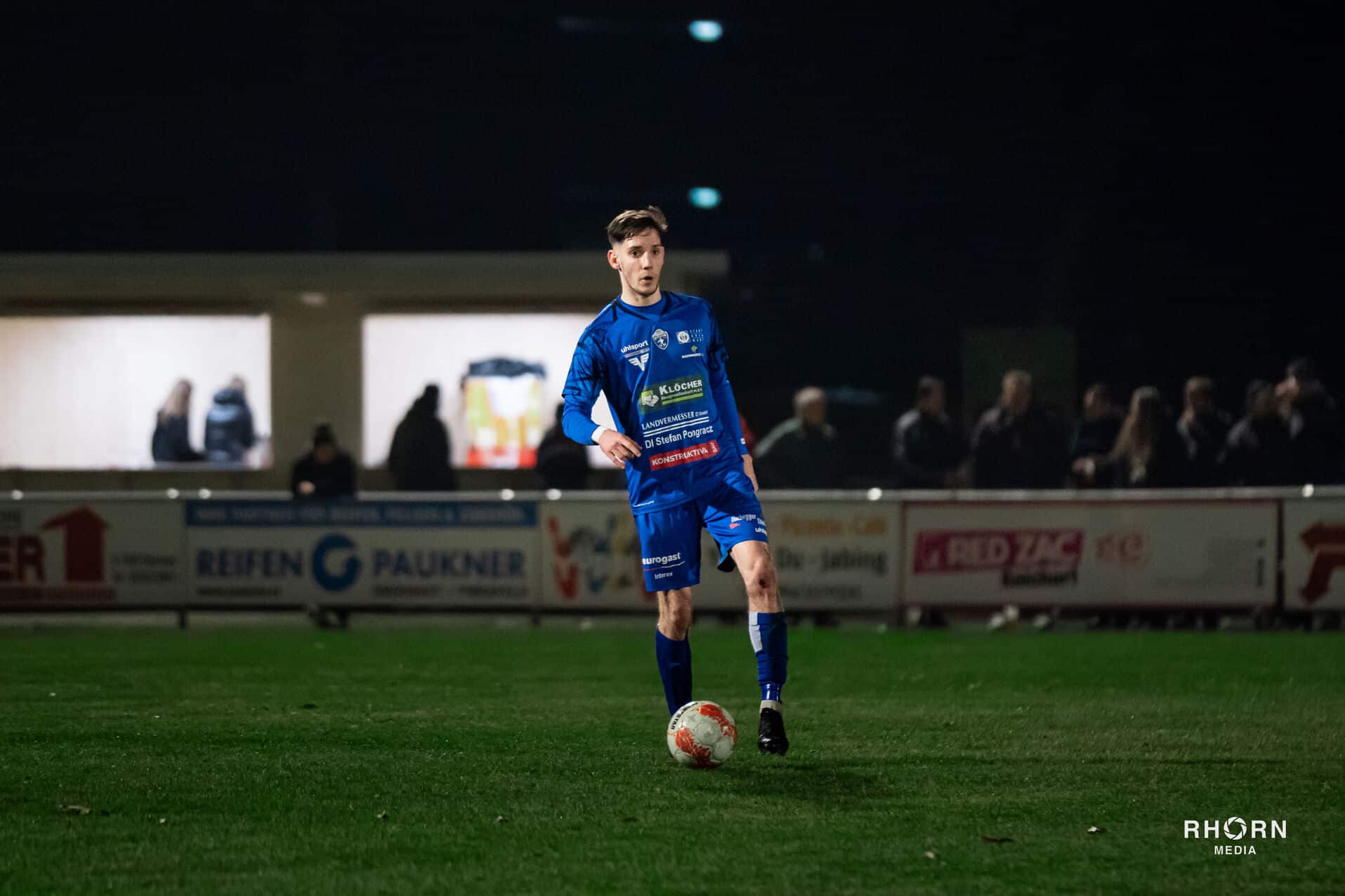Fußballspieler in blauem Trikot kontrolliert den Ball auf dem Spielfeld bei einem Nachtspiel.