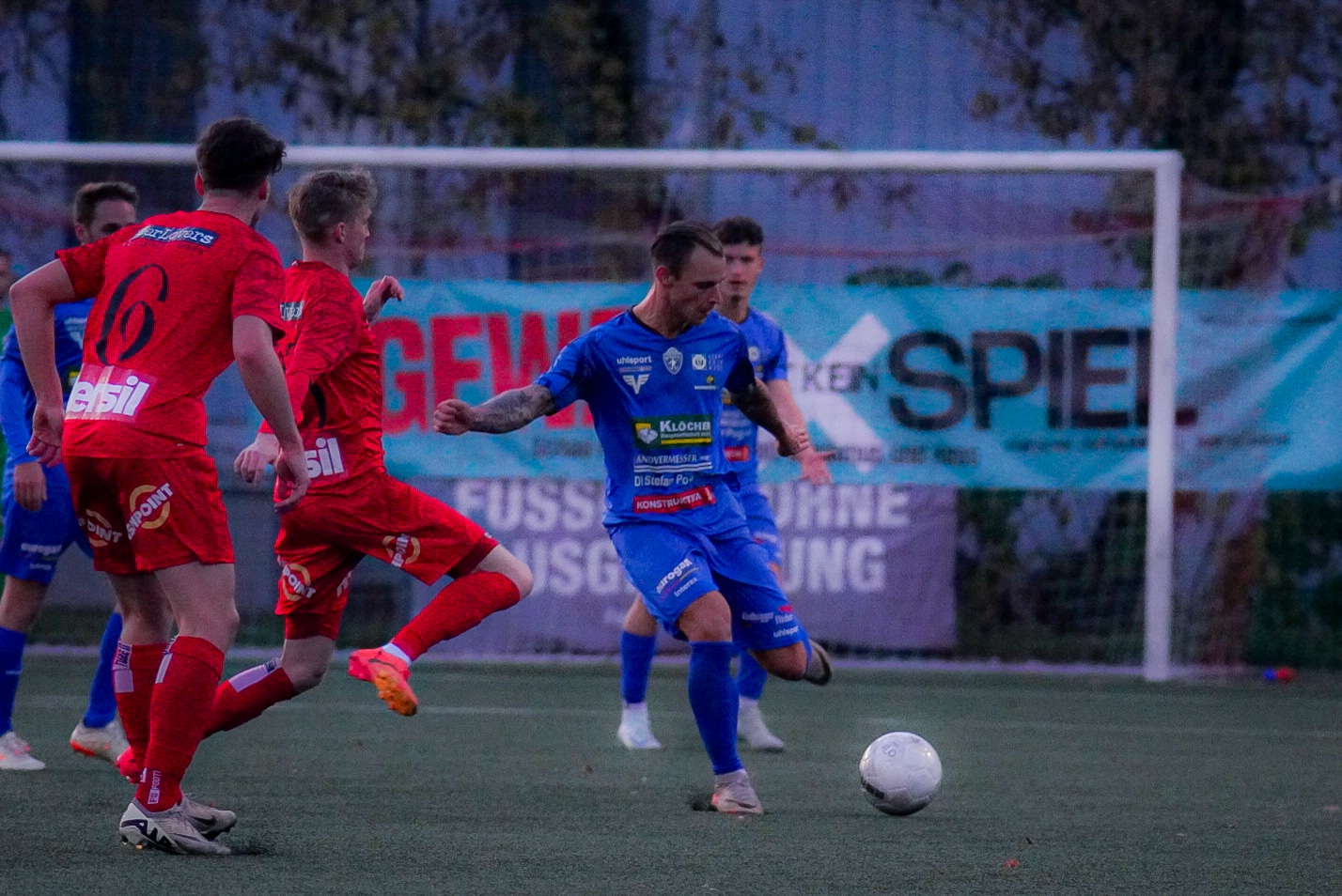 Fußballspiel in Aktion: Spieler in roten und blauen Trikots kämpfen um den Ball auf dem Spielfeld.