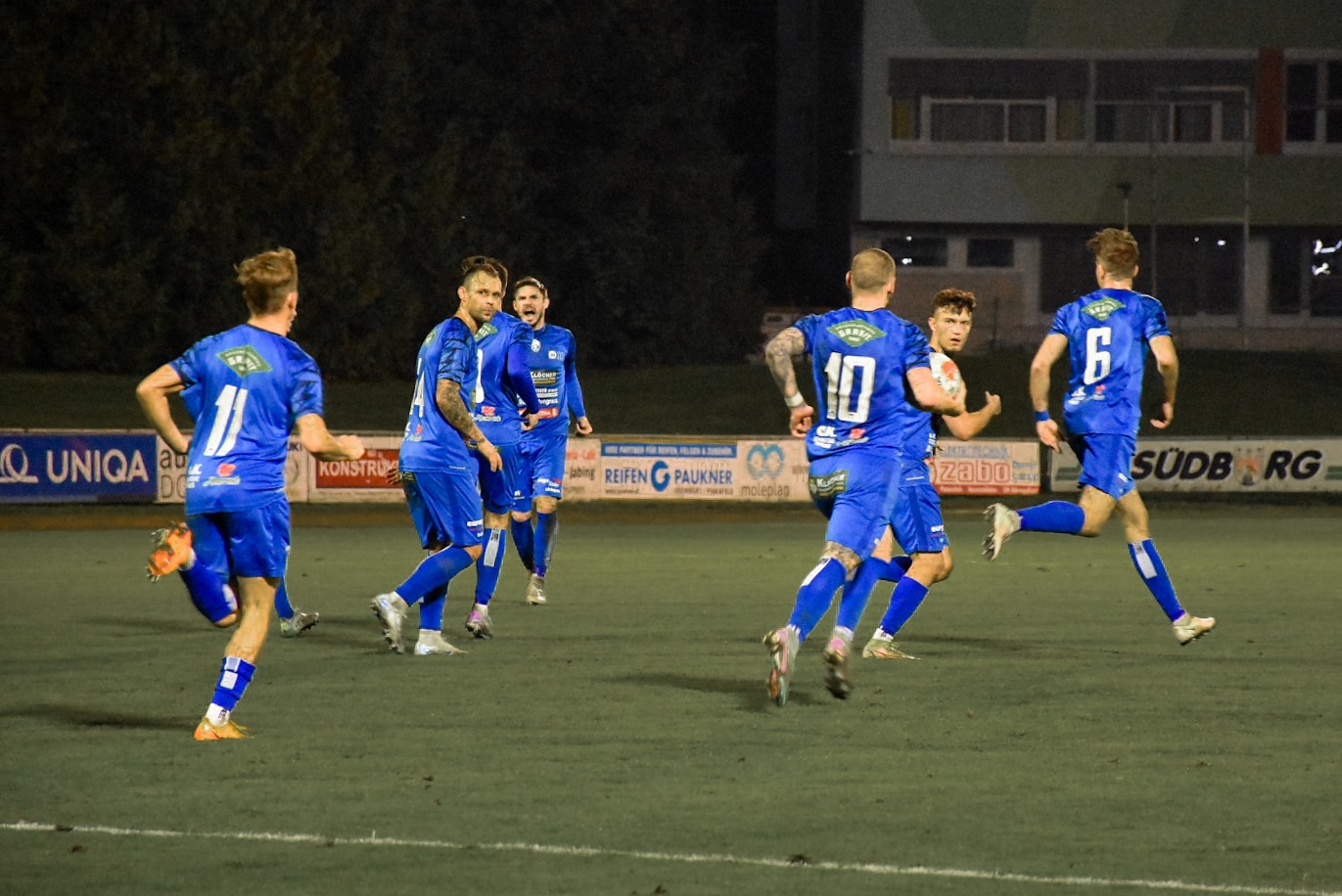 Fußballteam in blauen Trikots feiert auf dem Spielfeld während eines Abendspiels.