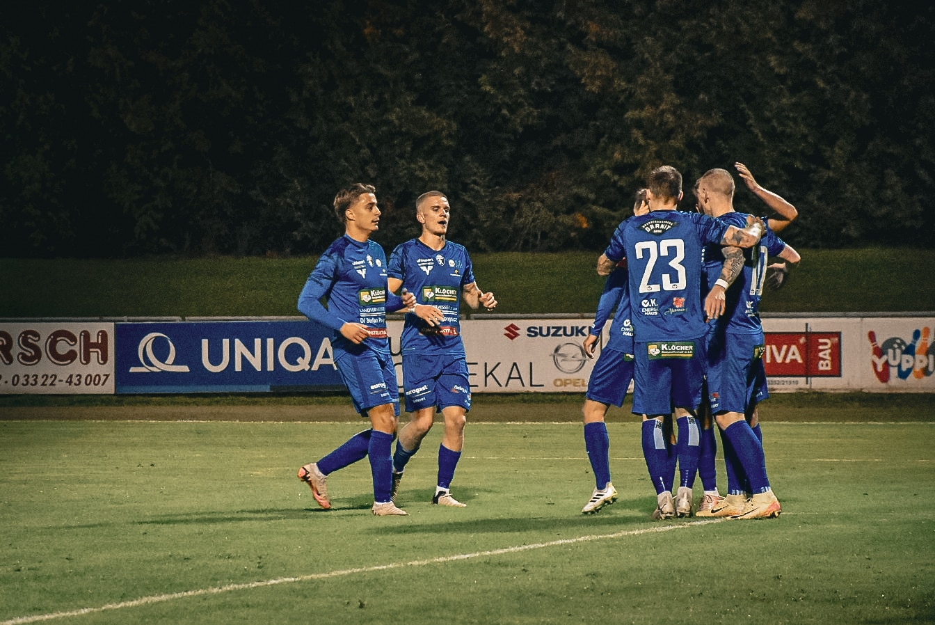 Fußballspieler feiern ein Tor in blauen Trikots auf dem Spielfeld, umgeben von Sponsorenwerbung.