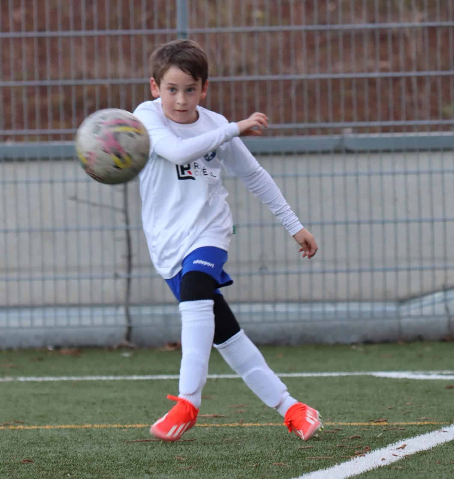 Junger Fußballspieler im weißen Trikot schießt einen Ball auf einem Kunstrasenplatz.