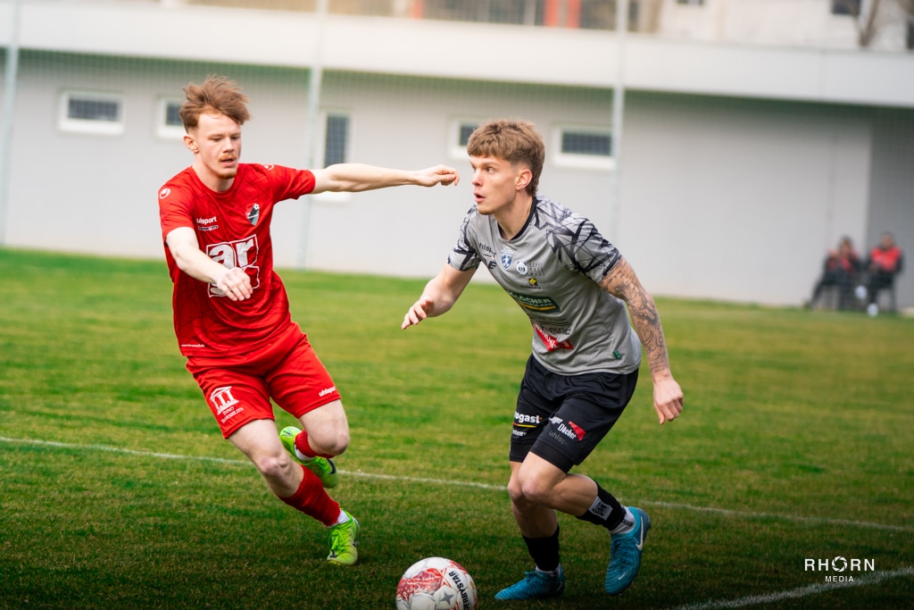 Zwei Fußballspieler kämpfen energisch um den Ball auf einem Spielfeld, ein Spieler im roten Trikot, der andere in Grau.