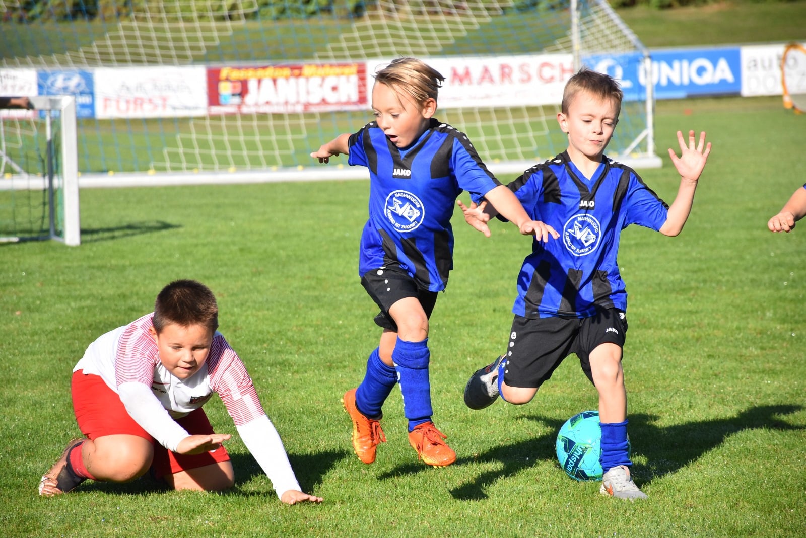 Kinder spielen Fußball auf grünem Rasen bei Sonnenschein, actionreiches Bild mit Jugendlichen in blauen Trikots.