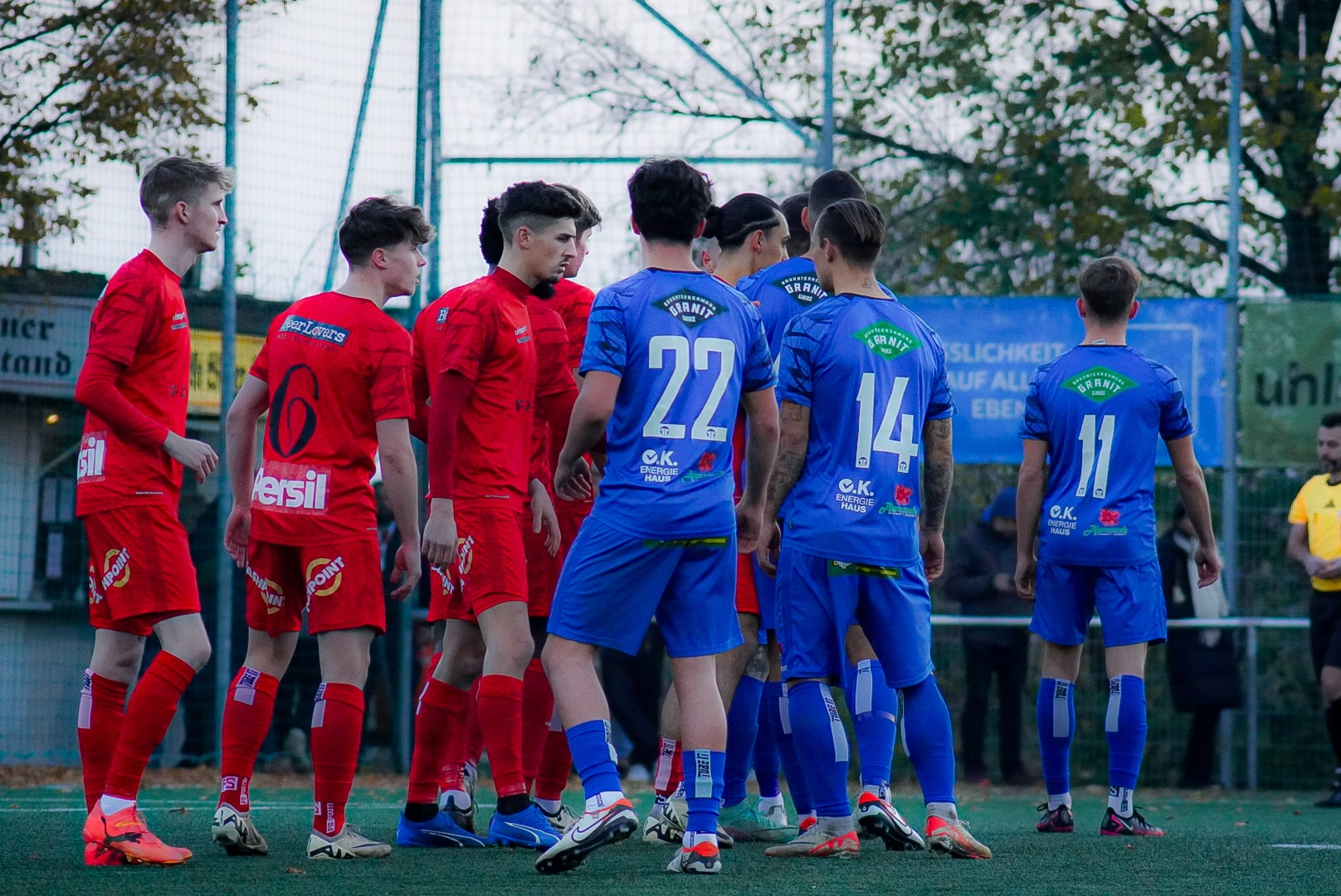 Spieler in roten und blauen Trikots stehen vor einem Fußballspiel auf dem Spielfeld; Teamgeist und Spannung.