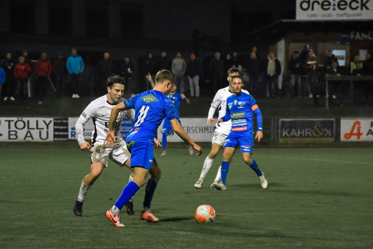 Fußballspiel bei Nacht: Zwei Teams im Duell um den Ball auf dem Spielfeld, Zuschauer im Hintergrund.
