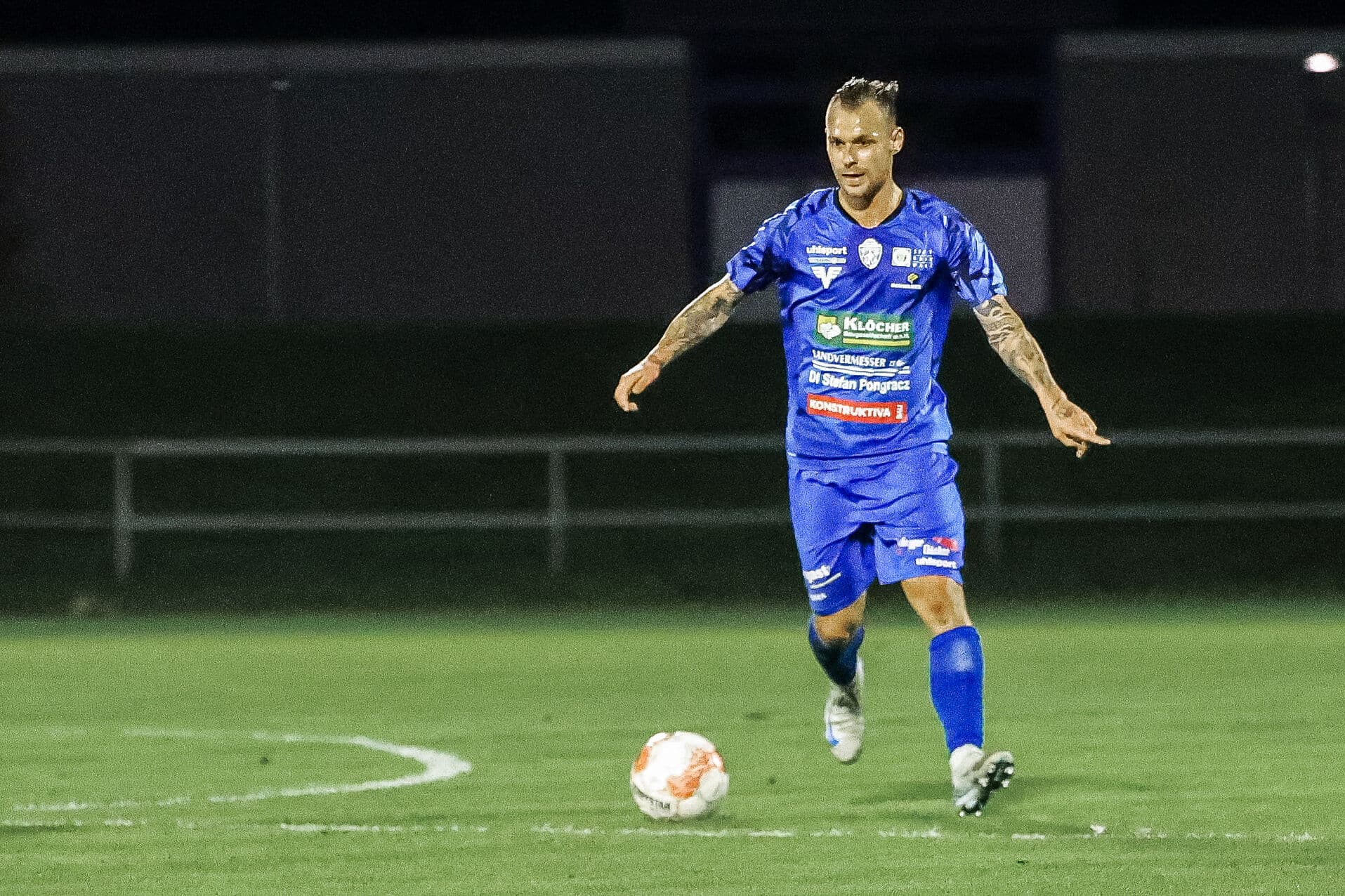 Fußballspieler in blauem Trikot dribbelt während eines Nachtspiels über das Feld.