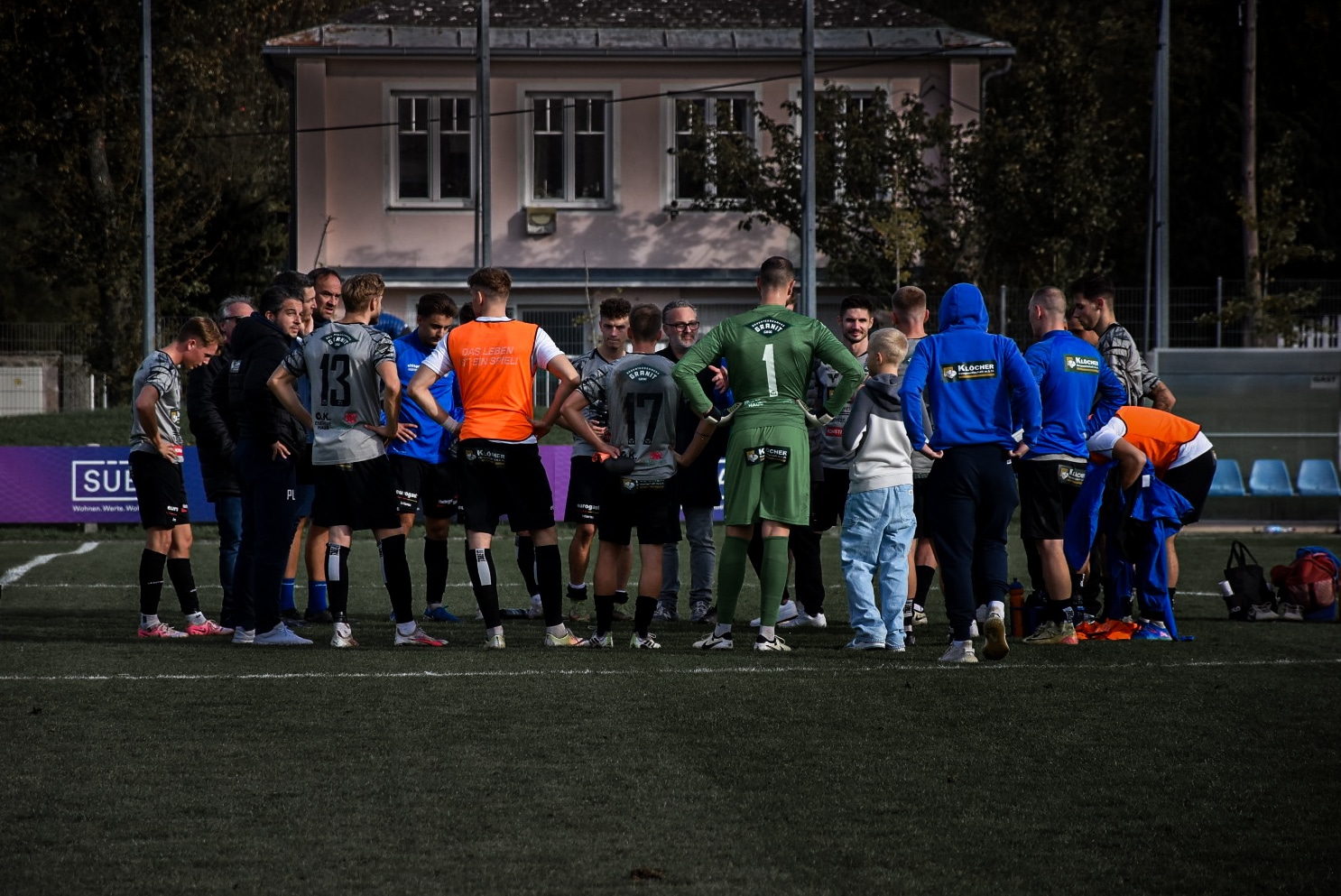Fußballmannschaft beim Teammeeting auf dem Spielfeld, Trainer motiviert Spieler in grauen und grünen Trikots vor einem Spiel.